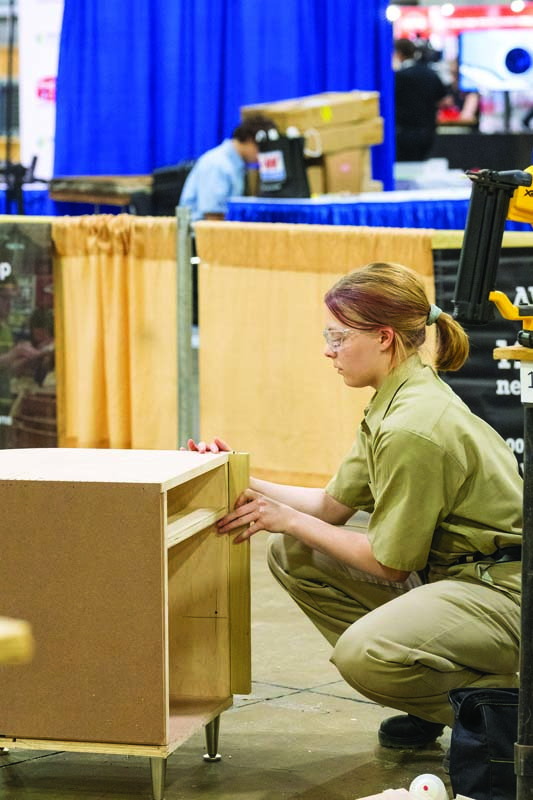 A person in a work uniform and safety glasses kneels to assemble a wooden cabinet at a trade show or workshop.