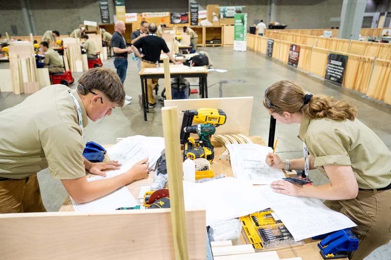Two individuals in uniform focus on blueprints at a workbench filled with tools and wood at a large indoor workshop event.